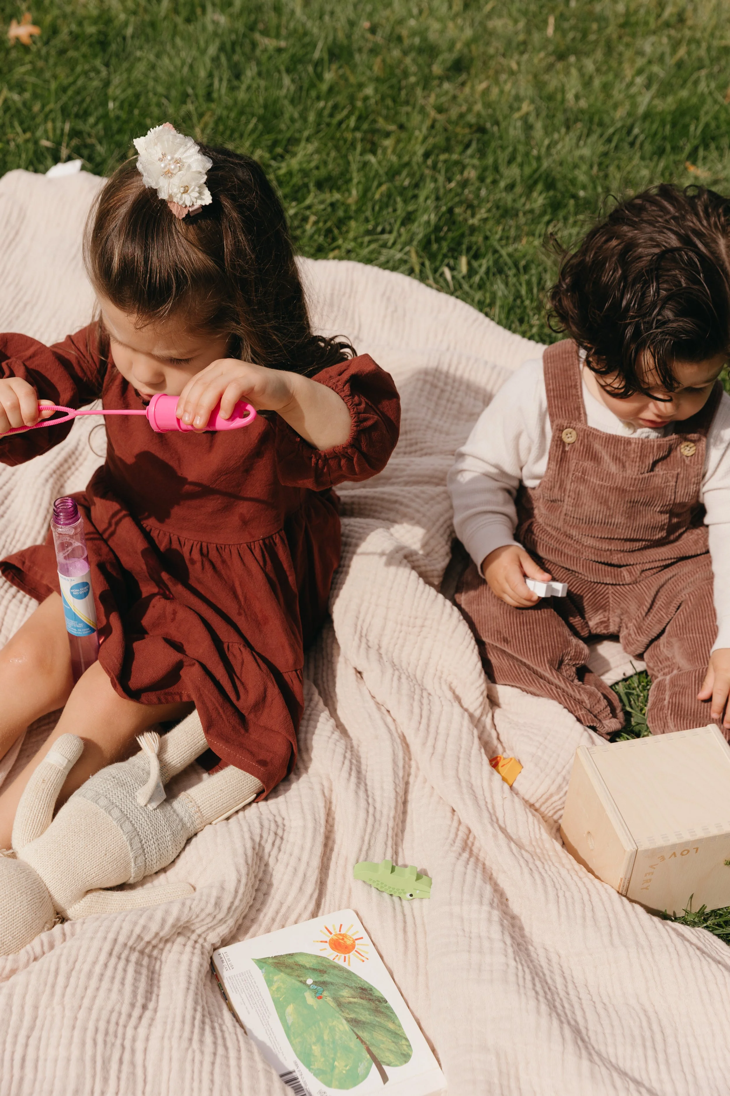 Two children sitting on a beige blanket outdoors on grass, playing and reading books.