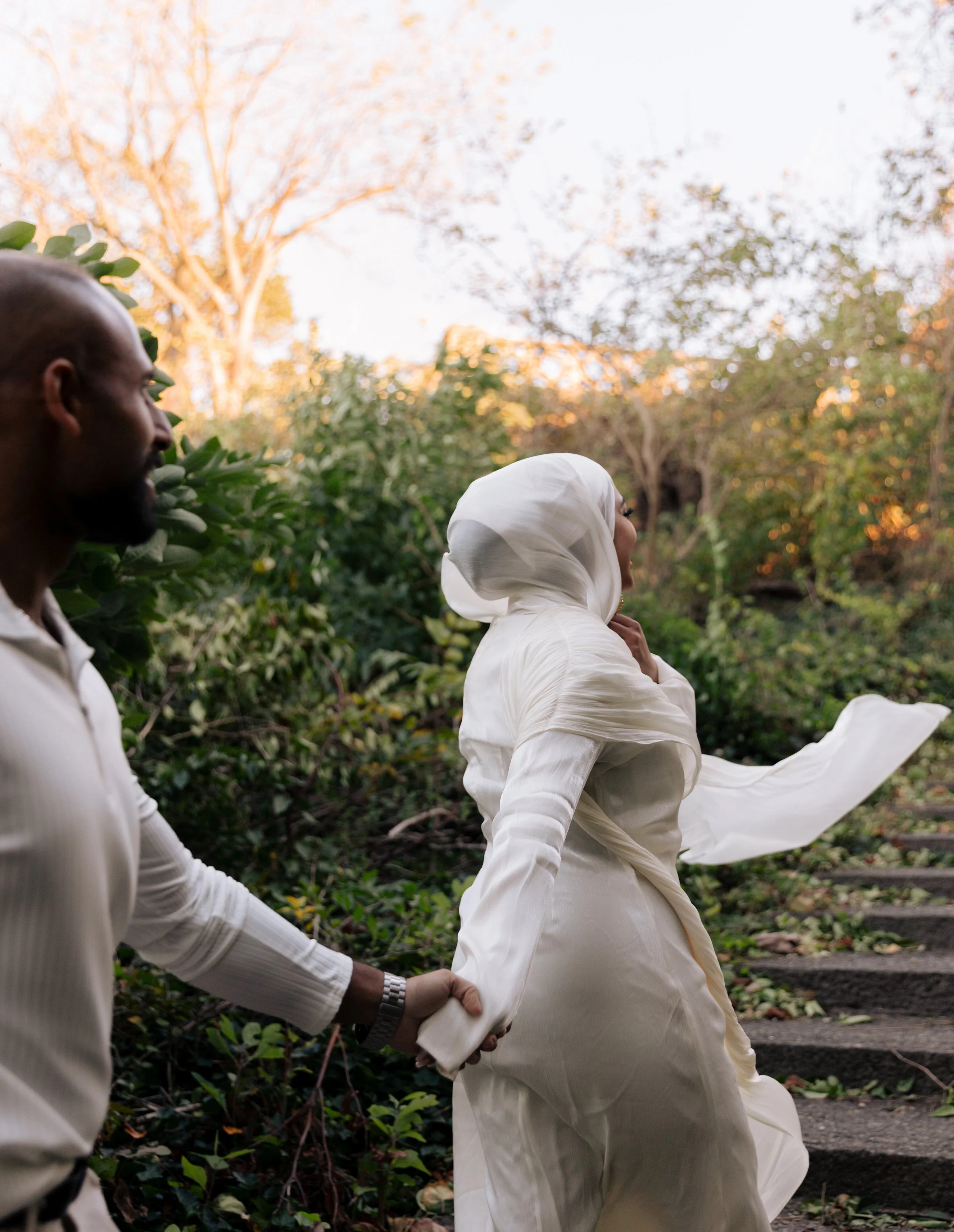 A man and a woman walk hand in hand outdoors in a garden or park during sunset. The woman is wearing a white hijab and white dress, and the man is dressed in a white shirt.
