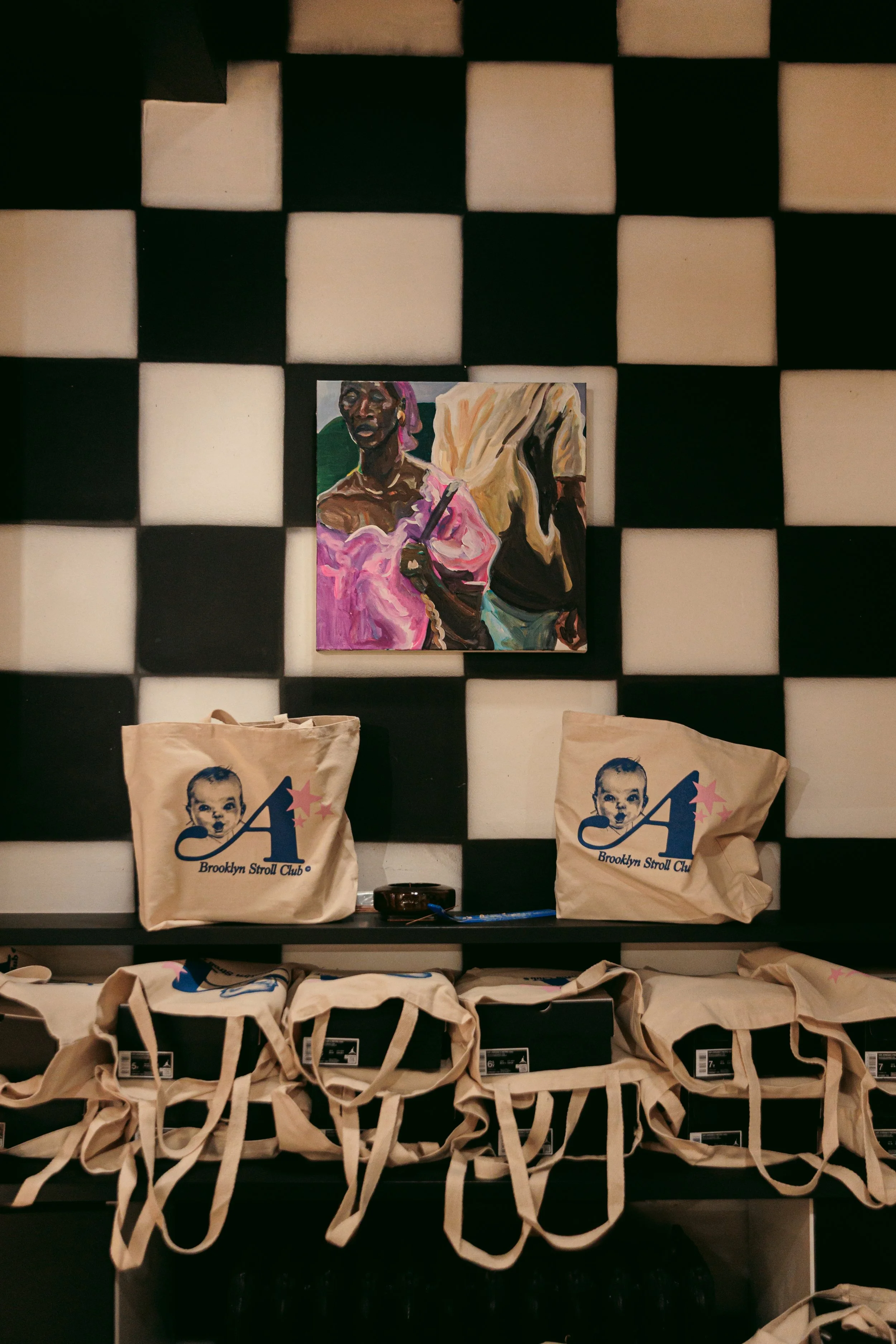 A display of reusable tote bags with a logo of a baby's face and the text 'Brooklyn Stroll Club,' set against a background of black and white checkered wall panels, with a colorful portrait painting hanging on the wall.