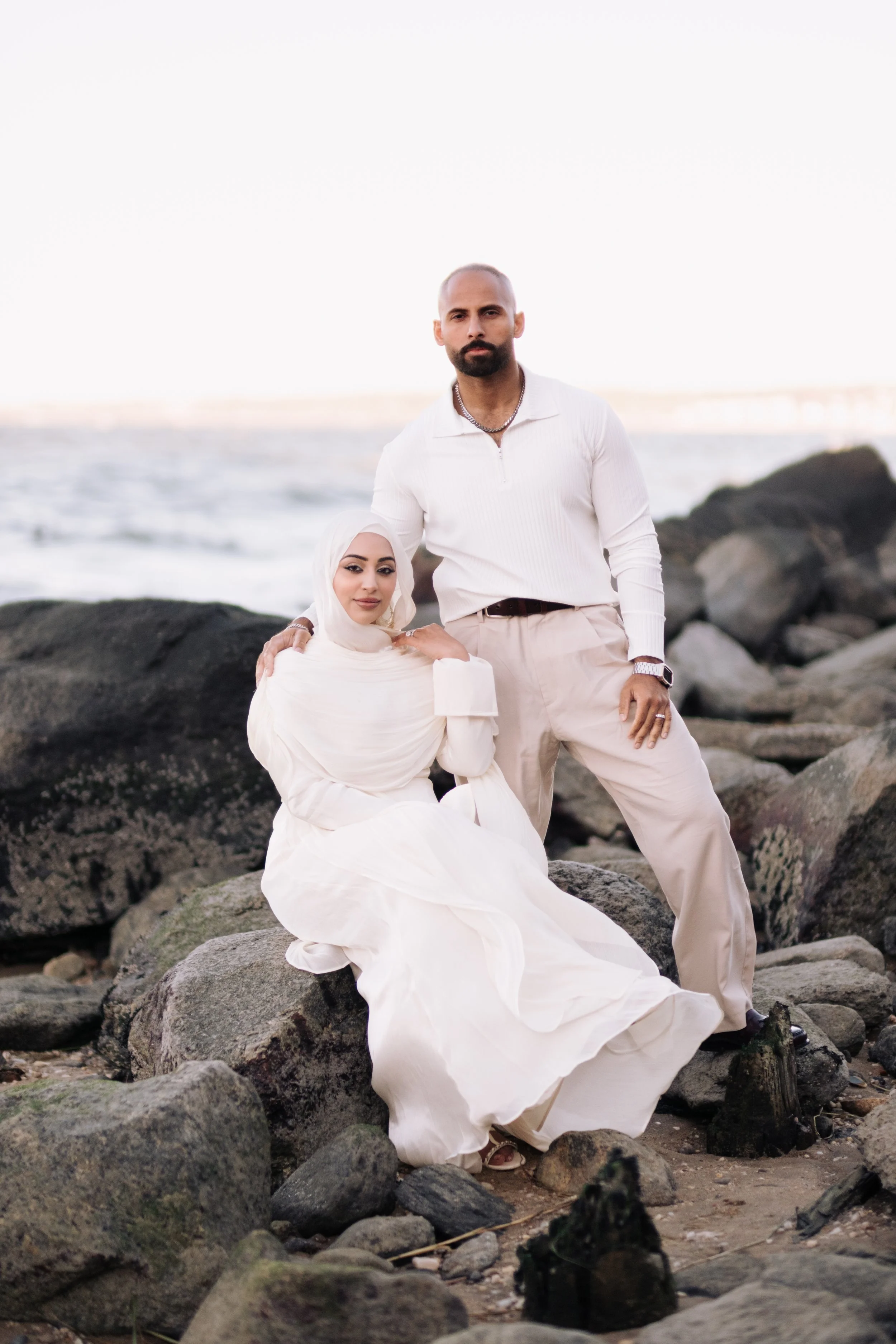 A man and woman on a rocky beach, dressed in white. The man stands with one hand on his hip, wearing a white shirt and beige pants, with a necklace and watch. The woman is seated on a rock, wearing a white dress and a white headscarf, smiling at the 