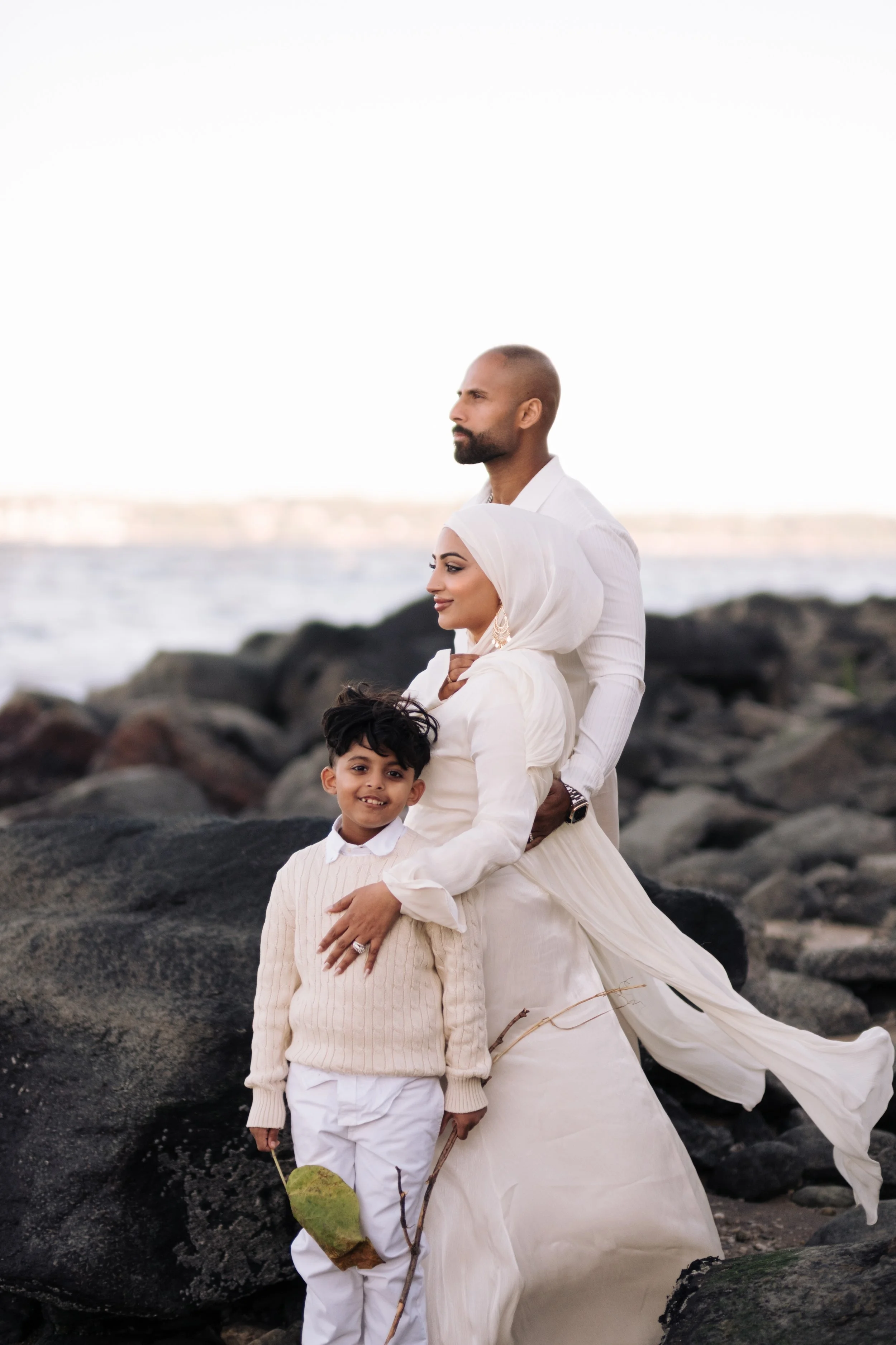 A family of three standing on rocks at the beach during sunset. The man is wearing a white shirt, the woman is wearing a white hijab and dress, and the boy is dressed in a beige sweater and white pants holding a leaf.