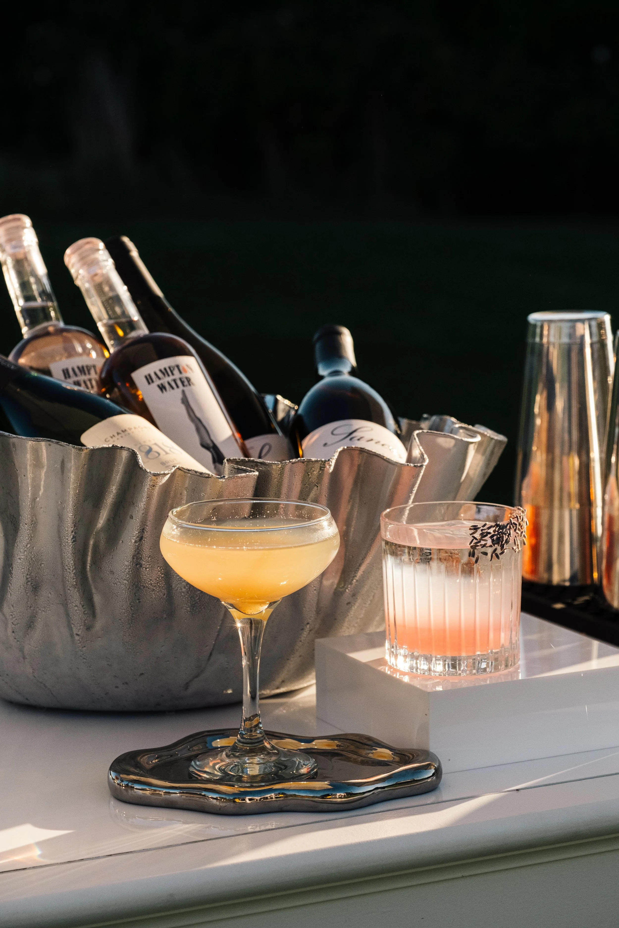 A silver ice bucket with bottled drinks labeled 'HAPPY WATER' and a champagne bottle inside, a cocktail glass with a yellow drink, and a pink beverage with black sprinkles on a white surface, against a dark background.