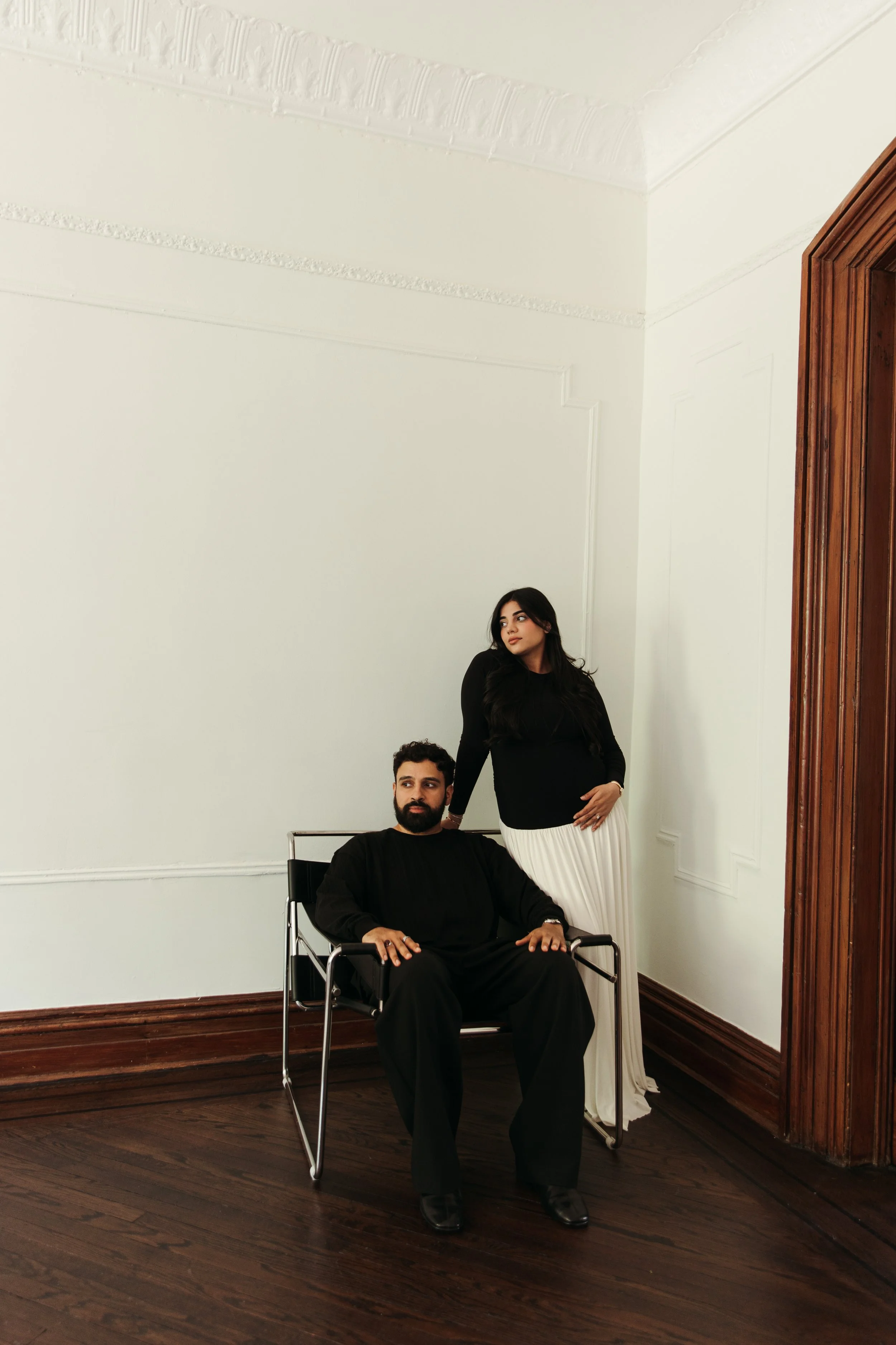 A man with a beard and a woman with long dark hair in a black top and white skirt in a room with white walls and dark wooden floor. The man is sitting in a wheelchair, and the woman is standing next to him resting her hand on his shoulder.