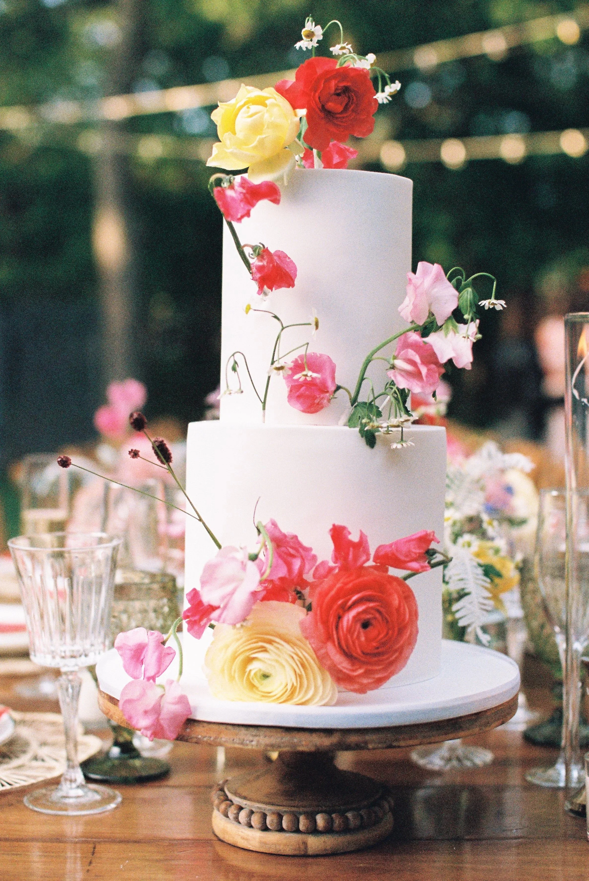 A multi-tiered wedding cake decorated with colorful fresh flowers, including roses and other blossoms, placed on a wooden stand at an outdoor celebration.