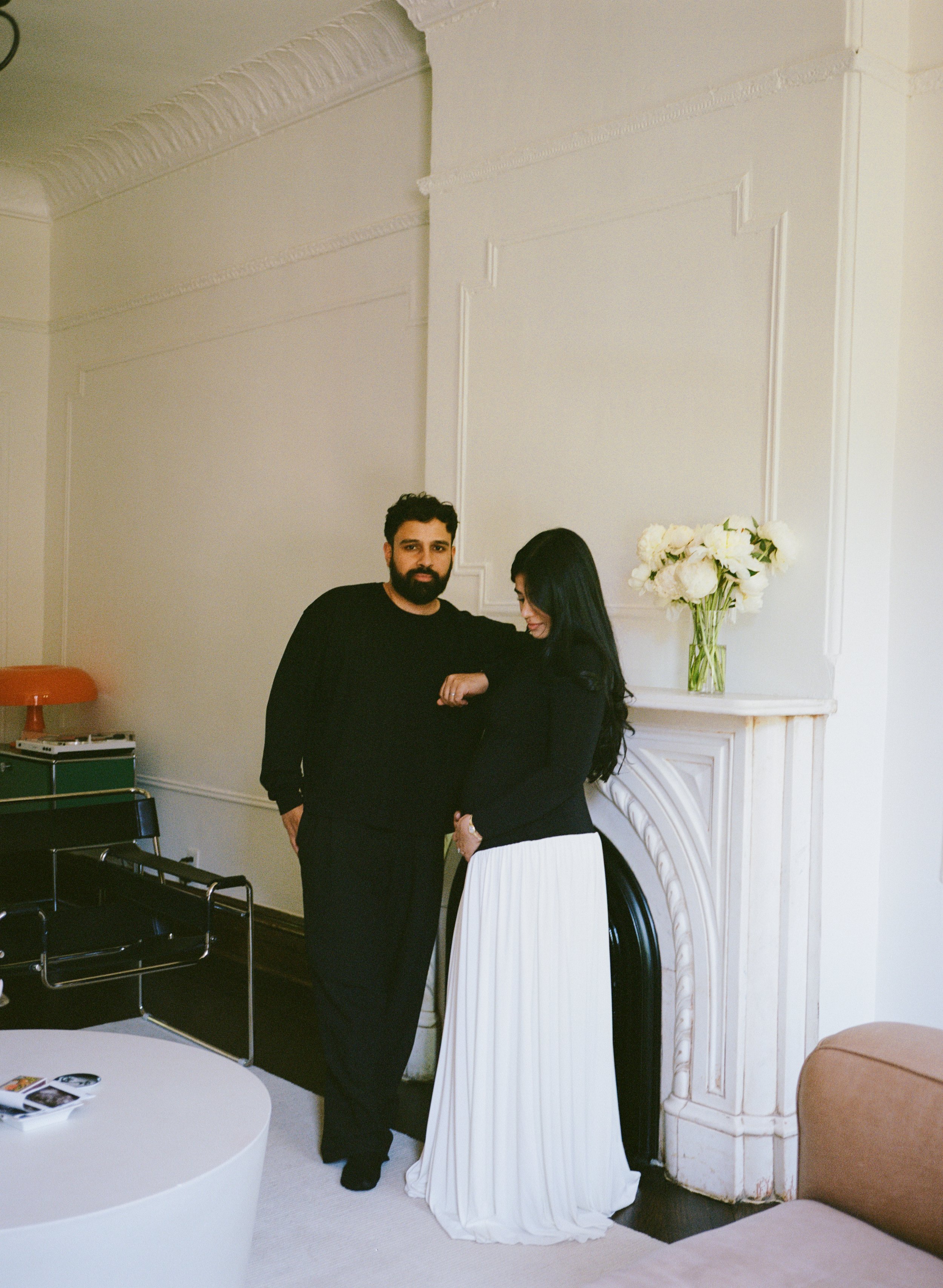 A man with dark hair and a beard wearing a black shirt and a woman with long dark hair wearing a black top and white skirt standing together in a room with beige walls, white decorative molding, a white fireplace, a vase of white flowers on the mantel, a round white coffee table with pictures, and an orange chair.