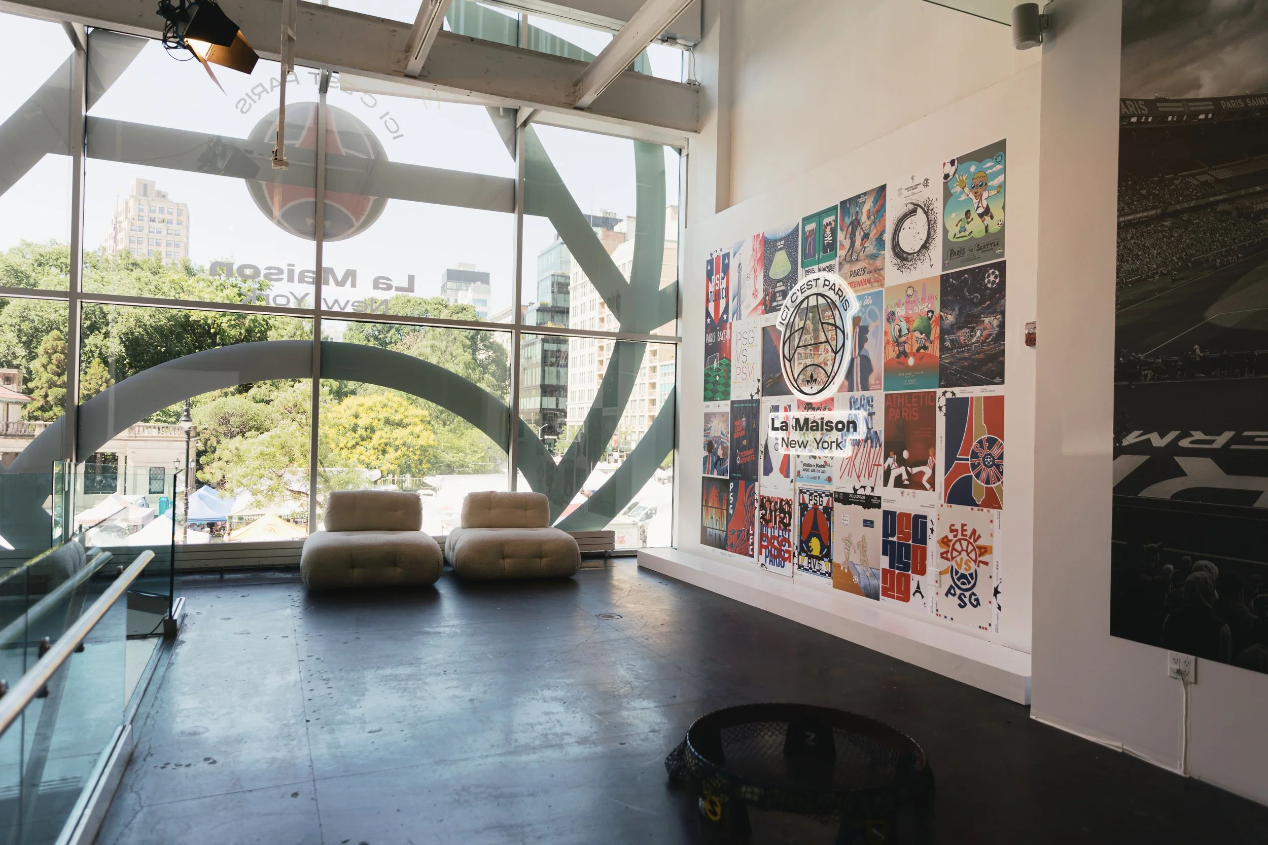 Interior of a modern art gallery with large glass windows, displaying colorful posters on the wall, two beige chairs, and a view of city buildings and trees outside.