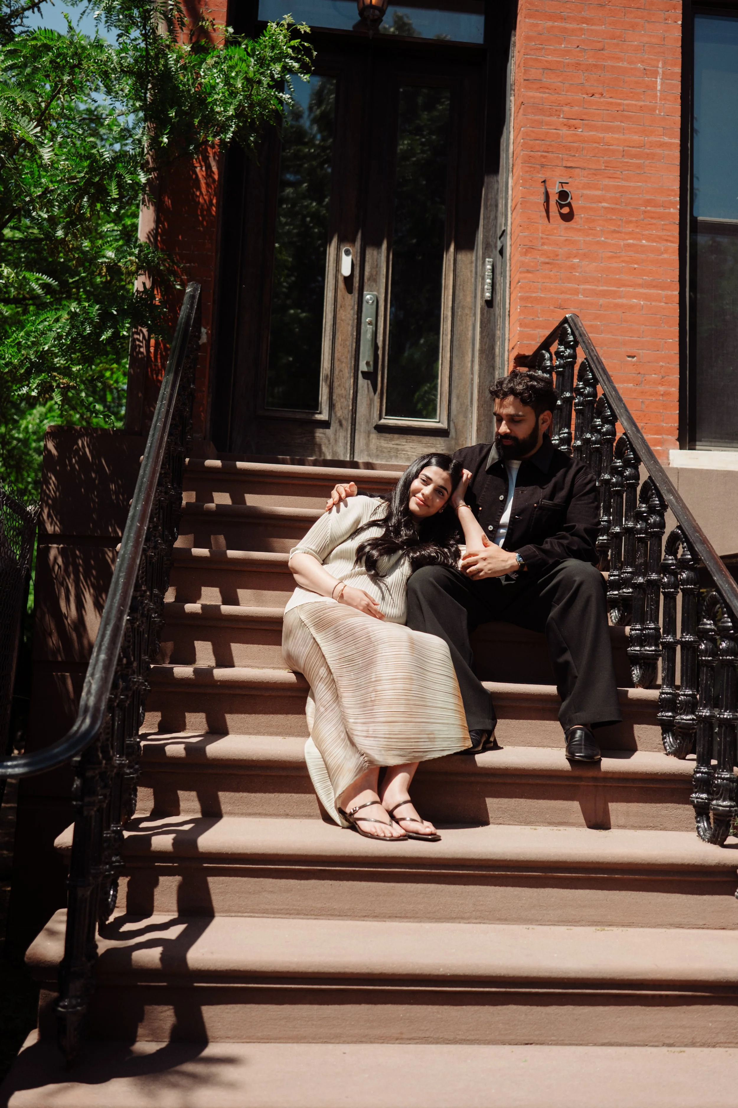 A young woman with long dark hair and a young man with short dark hair and a beard sitting on brownstone steps outside a brick building with large glass windows, surrounded by green trees.
