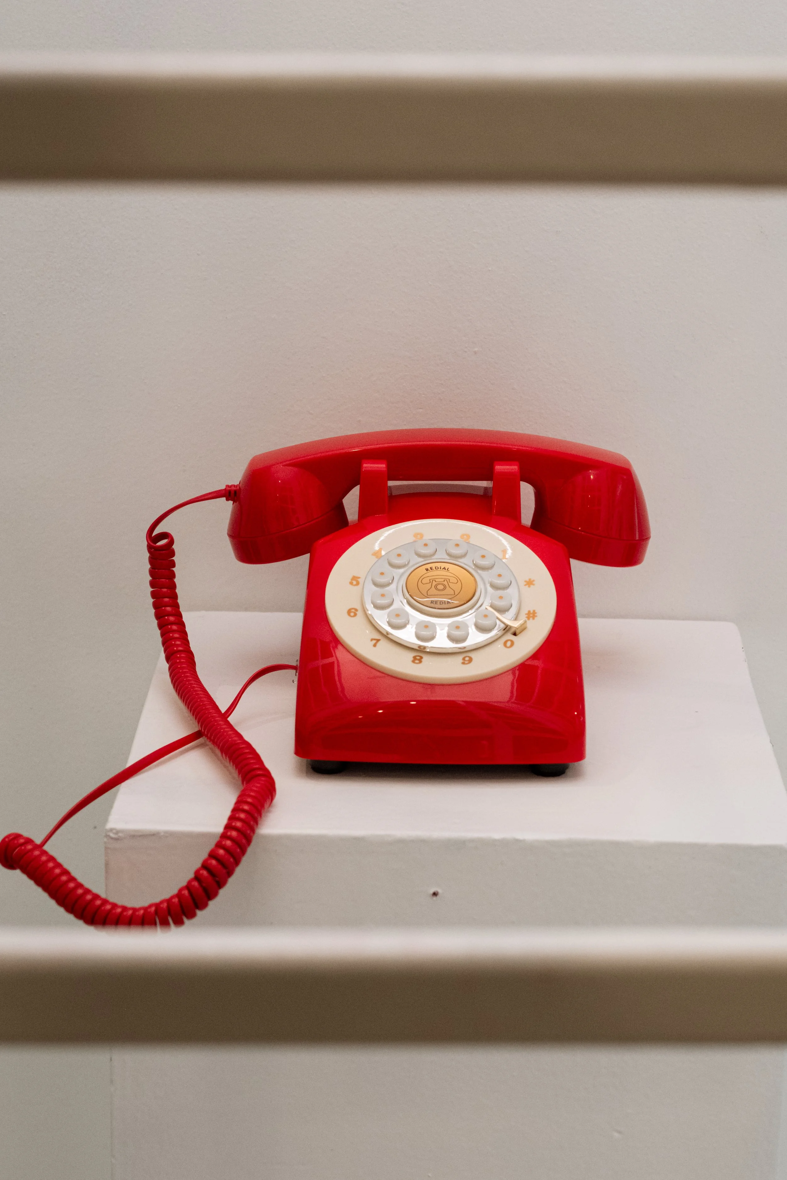 Red vintage rotary telephone with coiled cord displayed on a white pedestal against a beige wall.