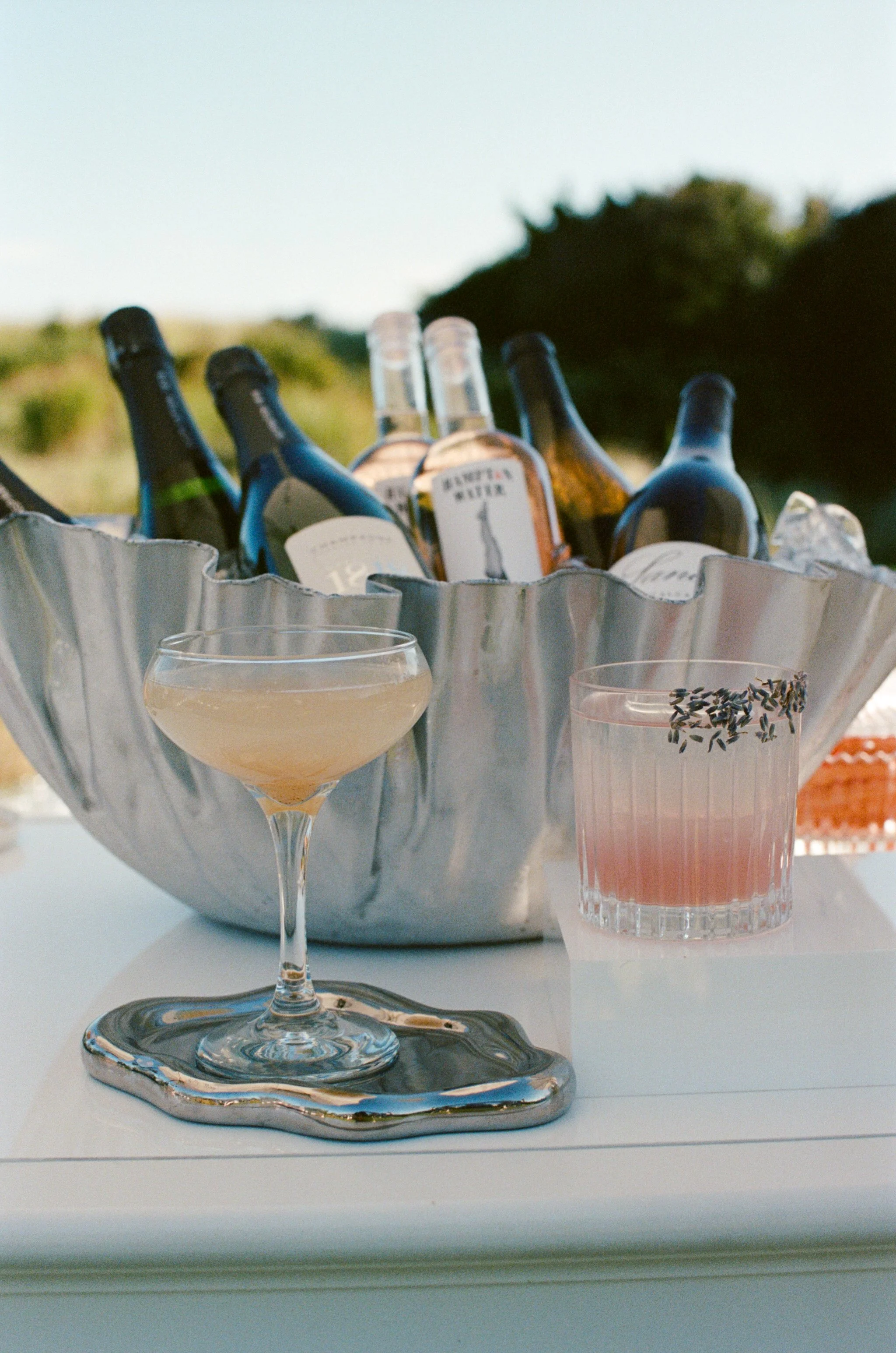 A silver bowl filled with bottles of alcohol, a glass with a light-colored drink and a sprig of lavender on top, a wine glass with a light beverage, and a silver tray with a candle, all set outdoors with trees and a sky in the background.