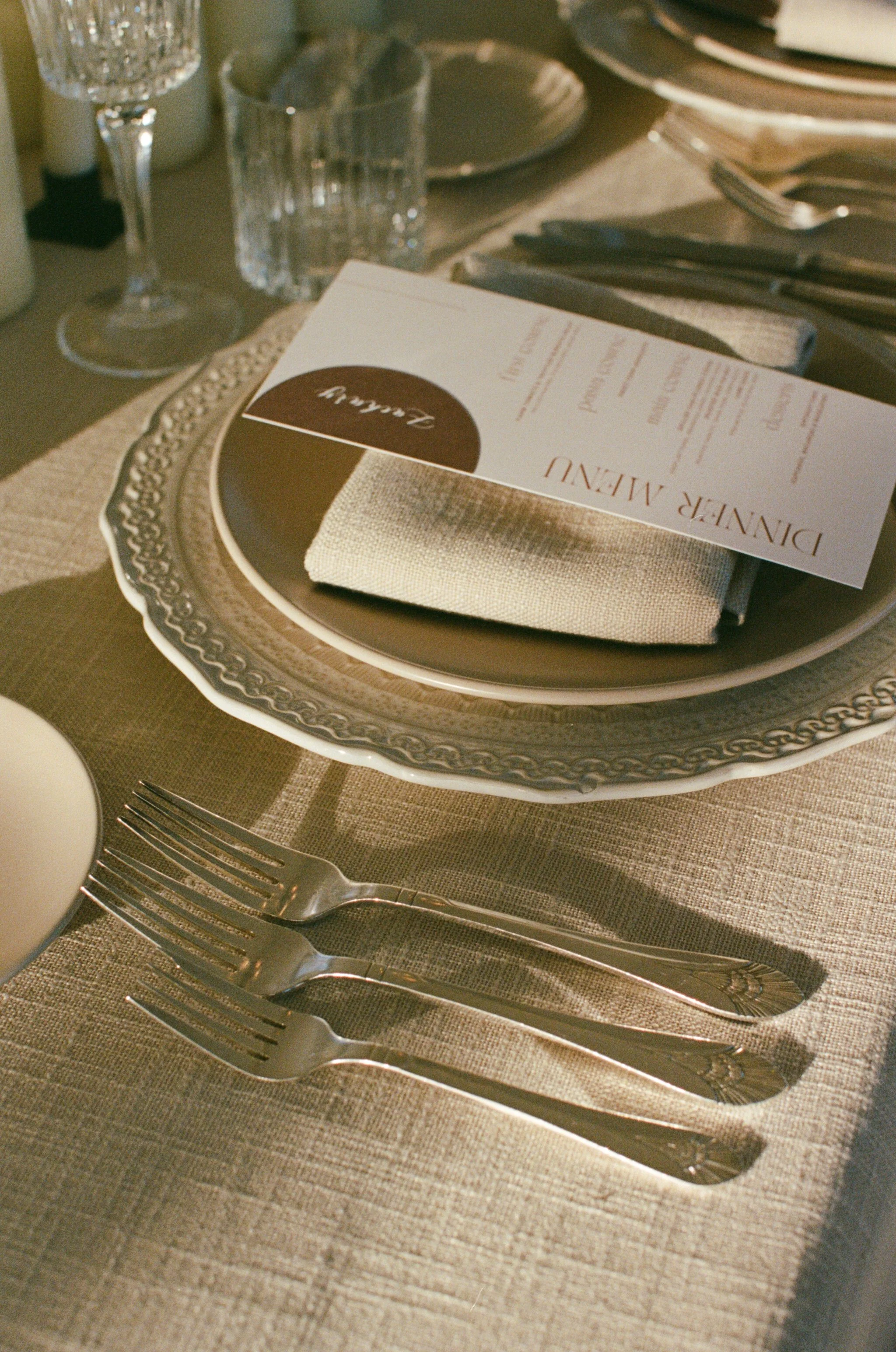 Elegant place setting with a decorative charger plate, a folded napkin, cutlery, a menu card, a glass, and a goblet on a table with beige cloth and striped placemat.