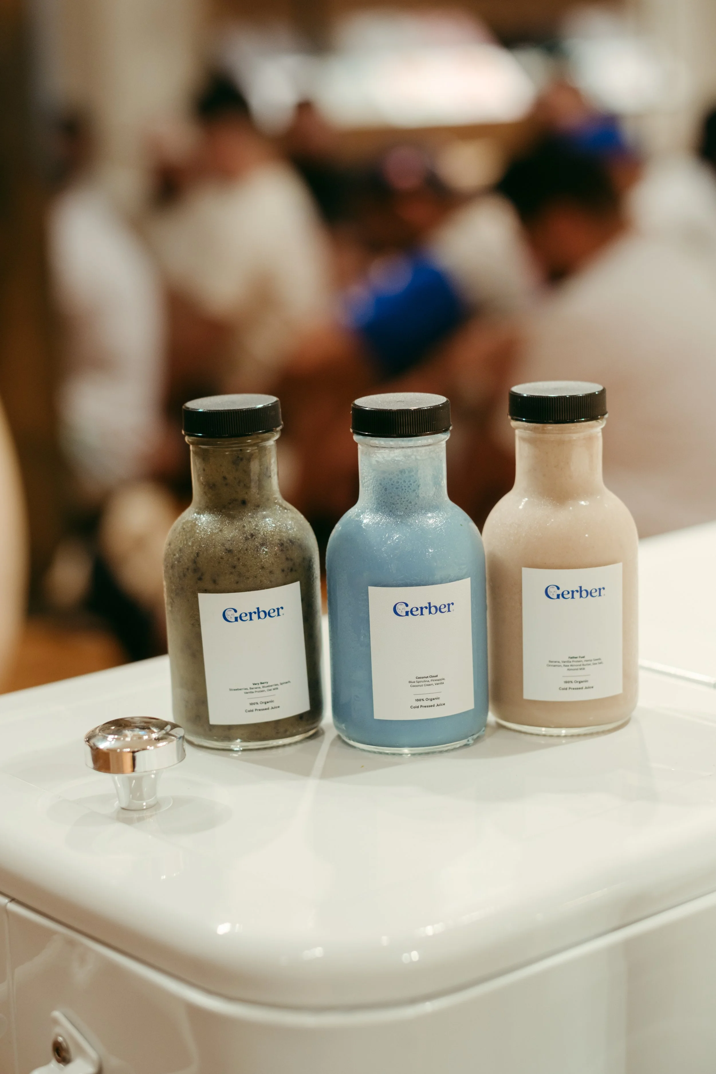 Three bottles of juice labeled Gerber on a white surface with a blurred group of people in the background.