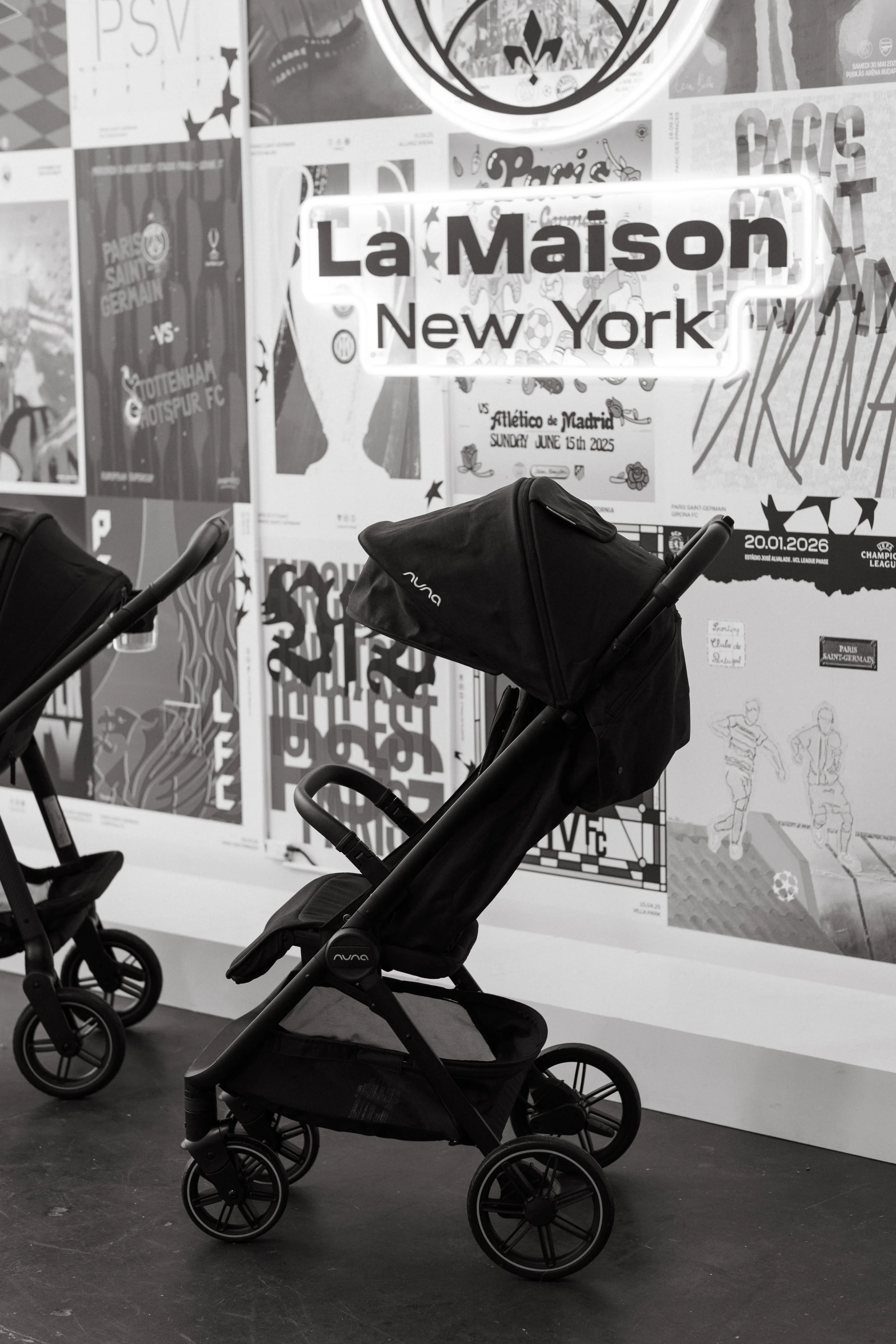 Black stroller with a fabric cover and large wheels, displayed in front of a wall decorated with various posters, including one for a soccer match between Paris Saint-Germain and Tottenham Hotspur FC.