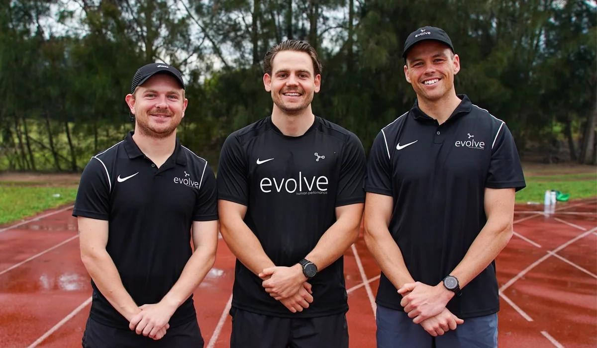 Three men standing on a running track outdoors, smiling at the camera, wearing black sports shirts with 'evolve' printed on them.
