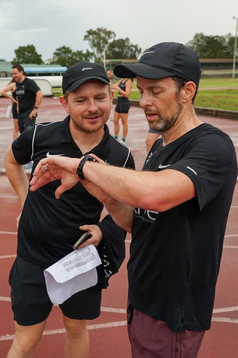 Two men in athletic clothing and caps looking at a sports watch on one man's wrist at an outdoor track, with other runners in the background.