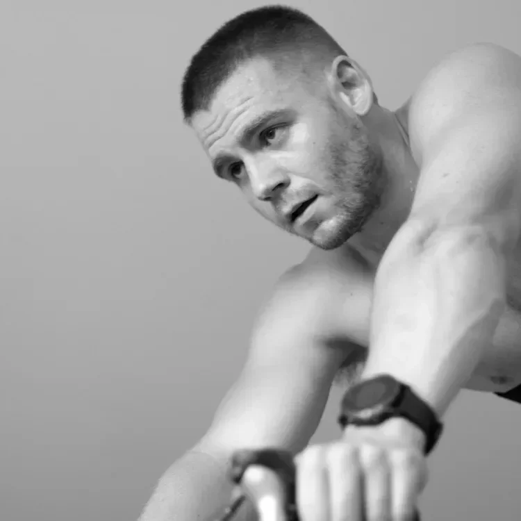 Black and white photo of a shirtless man with short hair and a beard, leaning forward with a focused expression, wearing a wristwatch, against a plain background.