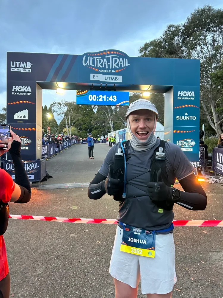Runner smiling at finish line of ultratrail race, wearing sports gear, bib number 3352, with the timing clock showing 21 minutes and 43 seconds.