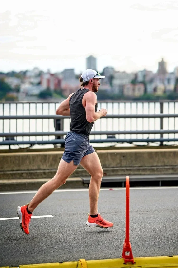 Man running on a street near a river with city buildings in the background, wearing athletic clothing and a hat.