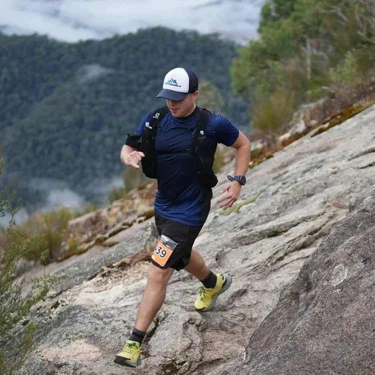 A man trail running on a rocky mountain path, wearing a blue shirt, black shorts, yellow running shoes, a cap, and a watch, with a mountain landscape in the background.