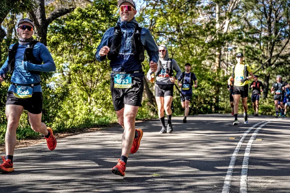 Group of runners participating in a marathon or race on a paved road surrounded by trees in a wooded area, wearing athletic gear and bib numbers.