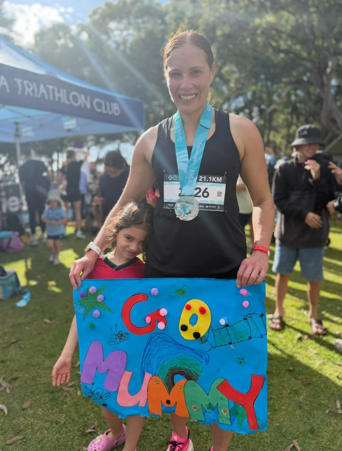 A woman with a race bib and medal, celebrating after a race, holding a colorful handmade sign that says 'Go Mum' with decorated letters and drawings, and a young girl standing beside her, in a park with people and a blue tent in the background.