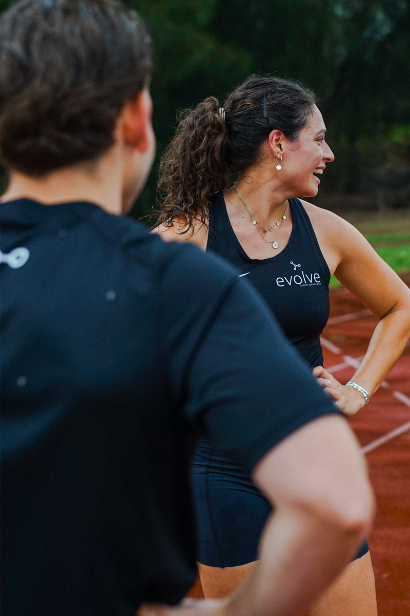 A woman smiling and talking outdoors with a man, on a running track, wearing workout clothes that say 'evolve.'