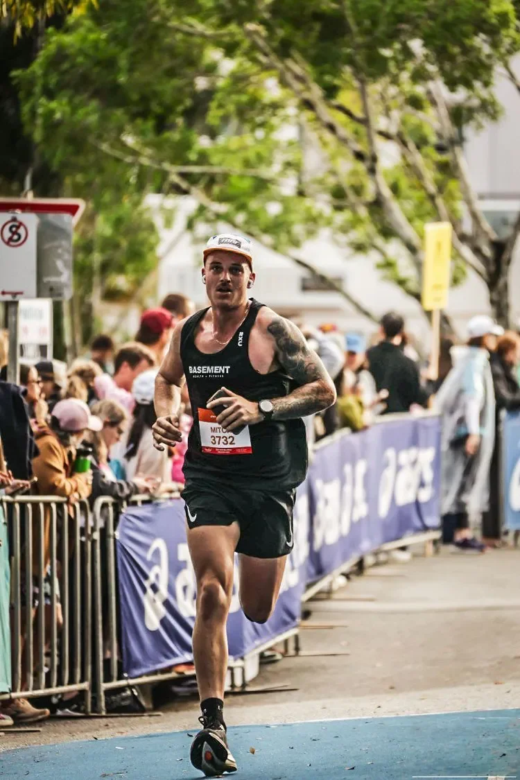 Male runner in black athletic gear running in a race on a city street, lined with spectators and trees.