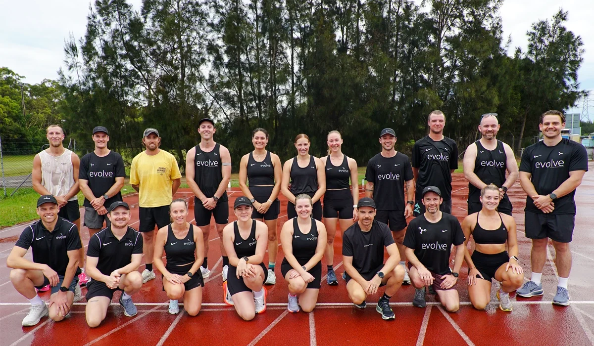 Group of athletes on a running track, wearing black athletic apparel with the word 'evolve' on some shirts, outdoors with trees in the background.