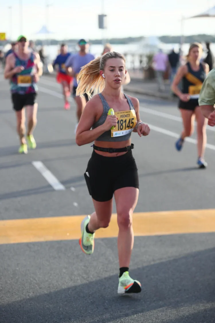 A young woman running in a marathon, wearing a gray sports bra and black shorts, with a yellow race bib numbered 18145, and earphones.