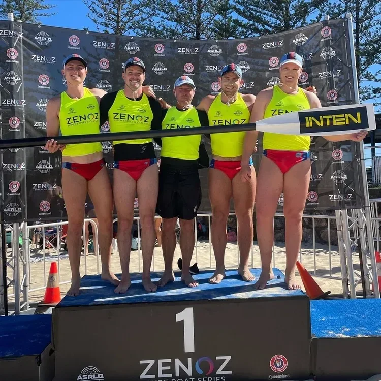 Group of five female surfers in yellow shirts and red shorts standing together on a winners' podium at a surfing competition, holding a giant paddle, with a black backdrop displaying event logos. They are smiling and celebrating after winning.