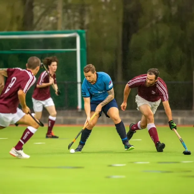 Hockey players competing for ball on a green field during a match under overcast sky.