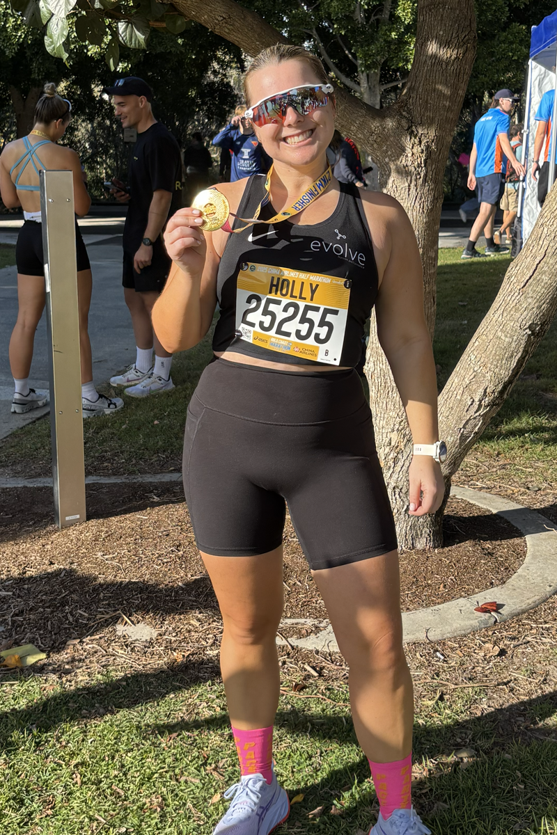 A woman in running gear holding a gold medal after finishing a race, smiling at the camera outdoors with trees and other runners in the background.