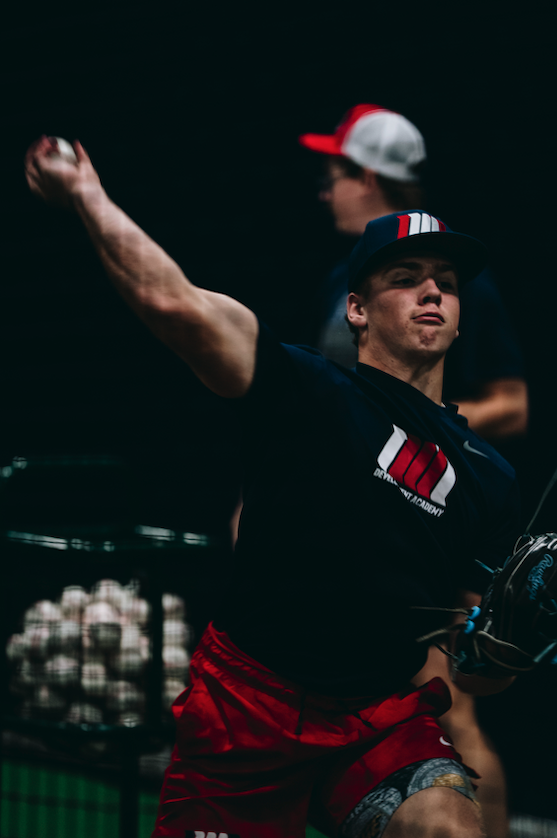 Development Academy pitcher mid throw with a bucket of baseballs behind him