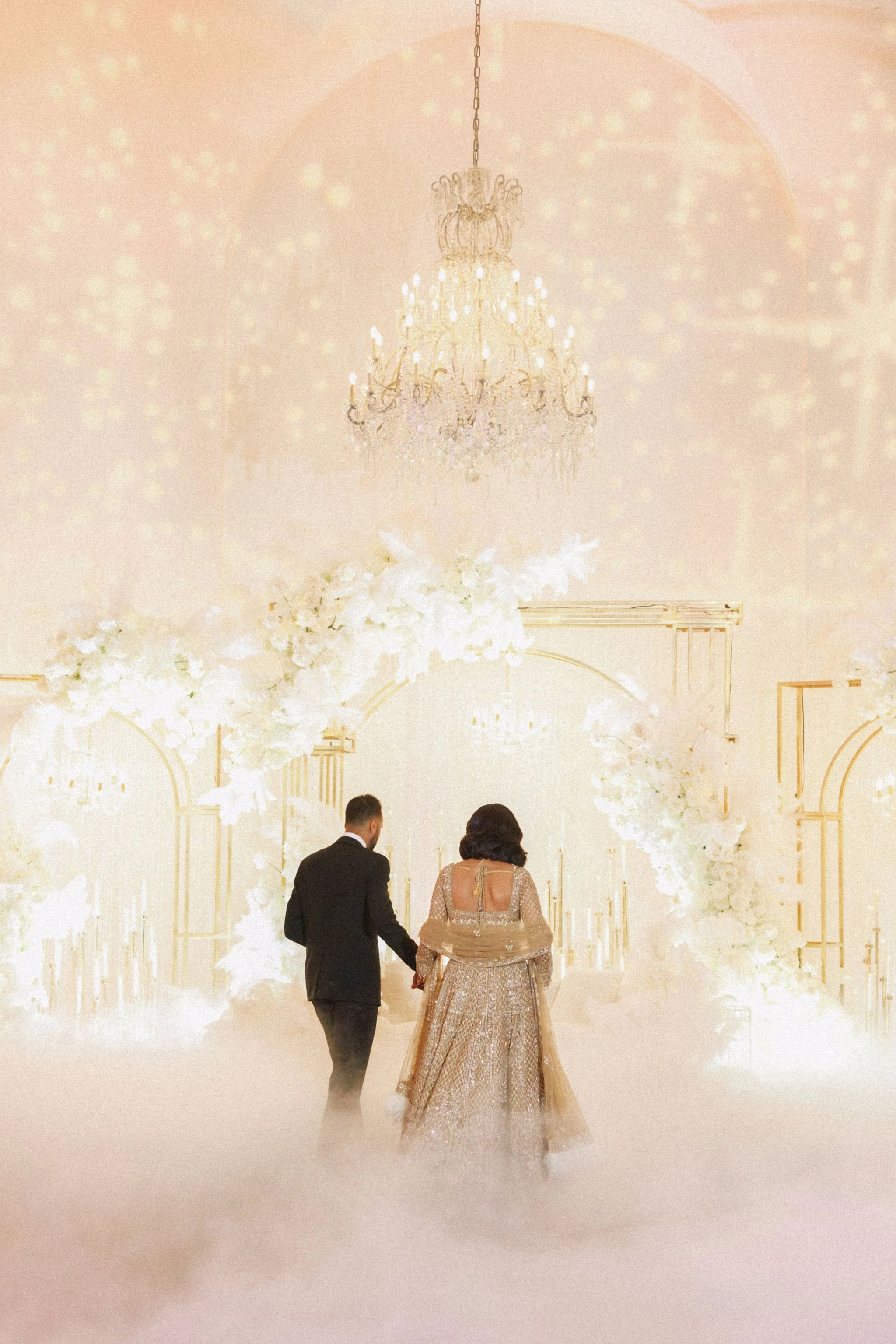 A bride and groom walking hand in hand through a wedding ceremony arch decorated with white flowers and candleholders, with a chandelier hanging above and soft, foggy lighting.