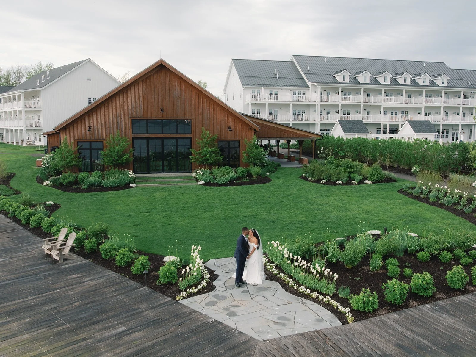 A bride and groom standing on a stone pathway in a lush garden with white flowers, holding hands and gazing at each other, with a wooden building and white residential buildings in the background.