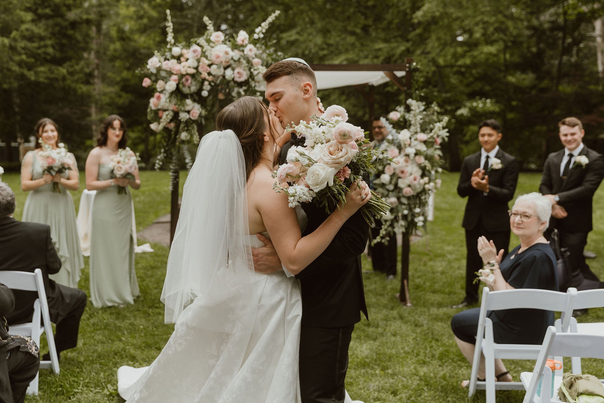 A bride and groom kiss during their outdoor wedding ceremony, surrounded by bridesmaids, groomsmen, and guests, with floral decorations and greenery in the background.