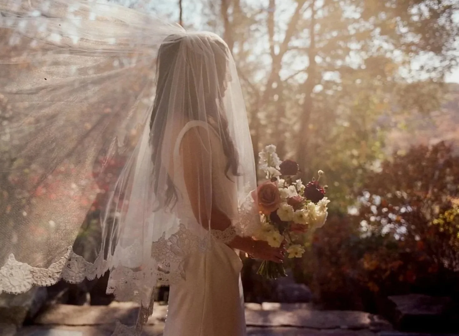 A bride wearing a lace wedding dress and veil, holding a bouquet of flowers, standing outdoors with autumn trees and sunlight in the background.