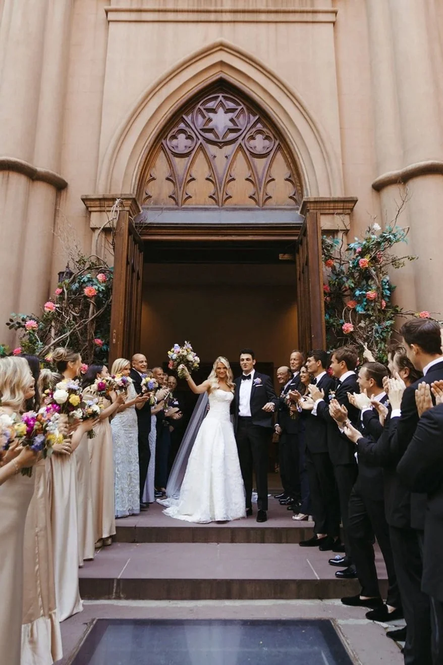 A bride and groom standing at the top of steps outside a church, surrounded by wedding guests clapping and holding flowers. The church has a large arched wooden door, floral decorations on either side, and intricate window details above.