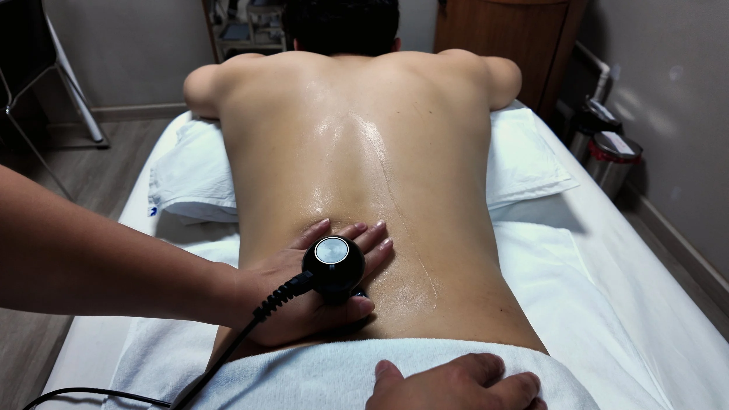 A person lying face down on a massage table while a therapist performs a massage in a dimly lit room with blue tones.