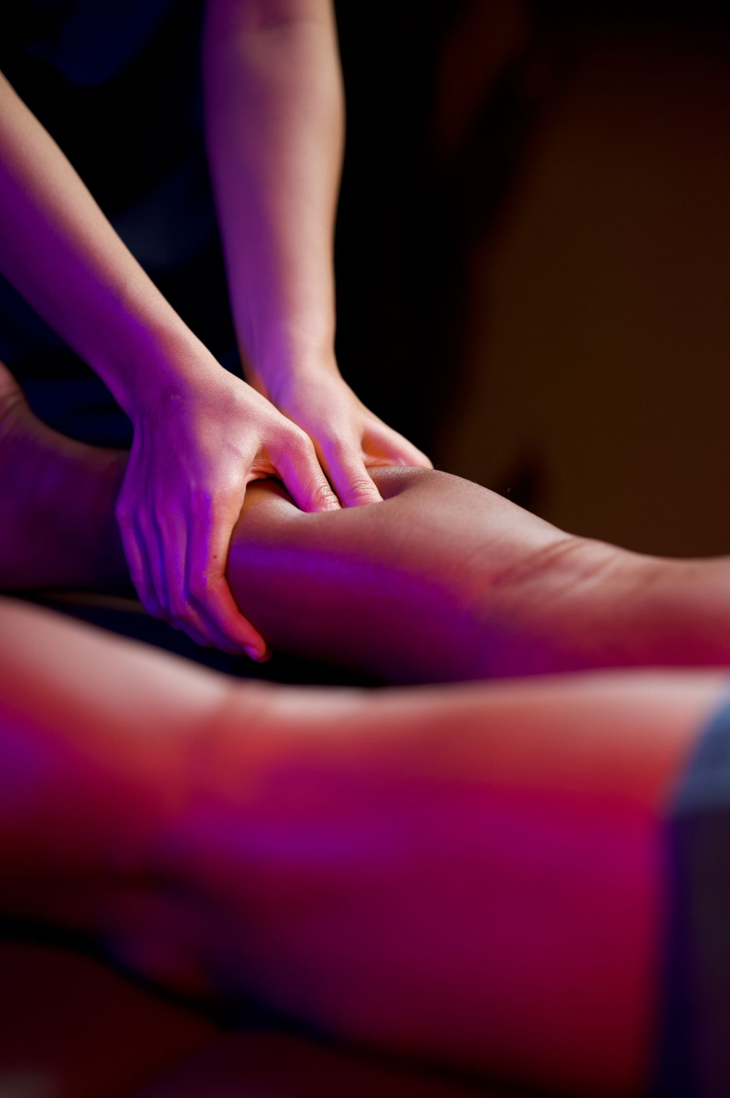 A person receiving a massage, with hands pressing on their leg in a dimly lit room with purple lighting.