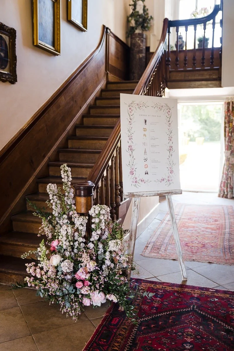 Flower arrangement with pink and white flowers next to an easel with a wedding order of the day sign, inside a house with wooden staircase and framed artwork on the wall.