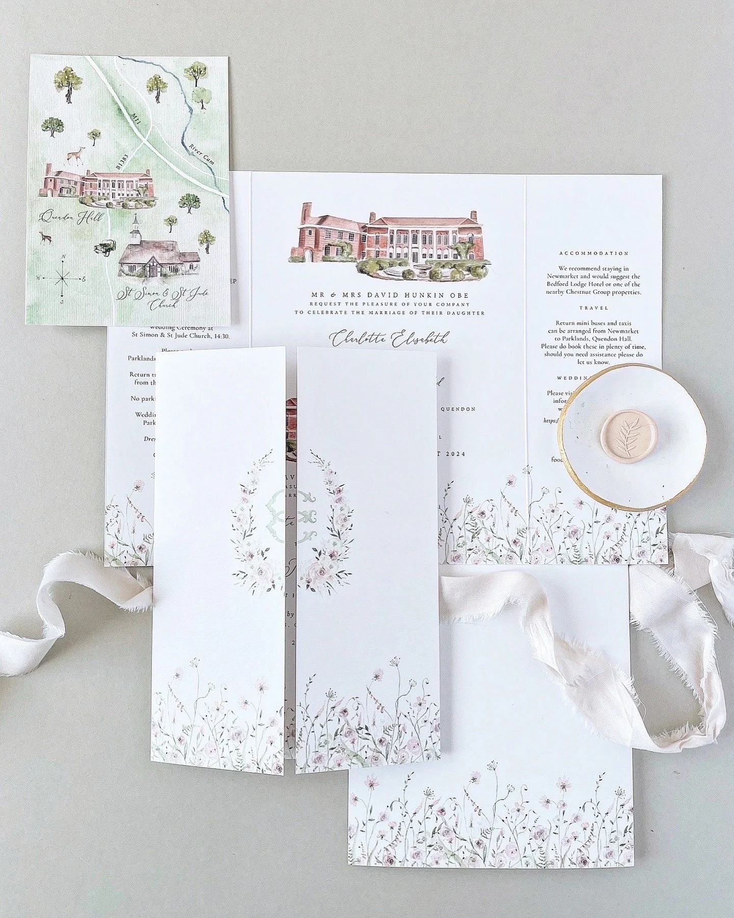 Wedding invitation suite with floral designs, map, and a small plate with a leaf emblem, arranged on a white surface with a white ribbon.