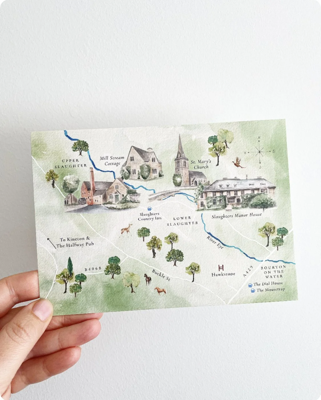 Hand holding a illustrated map of a rural village with trees, houses, a church, and a river running through, labeled with locations such as Slaughter Inn, St. Mary's Church, and Bourton on the Water.