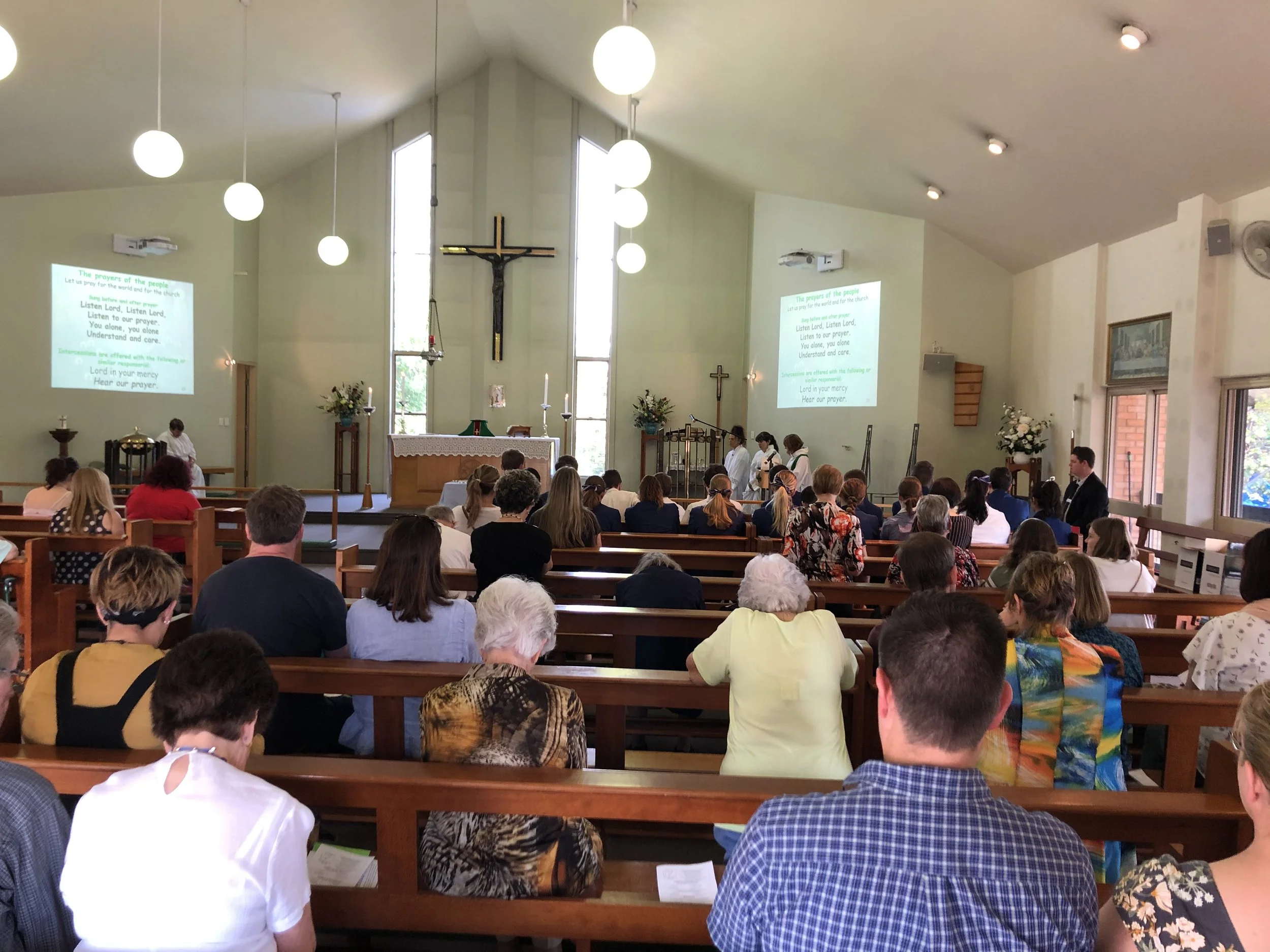 Inside a church during a service, with people seated in wooden pews facing the altar, which has a crucifix and candles. The church is well-lit with natural and artificial light, with hymn lyrics projected on two screens on either side of the altar.