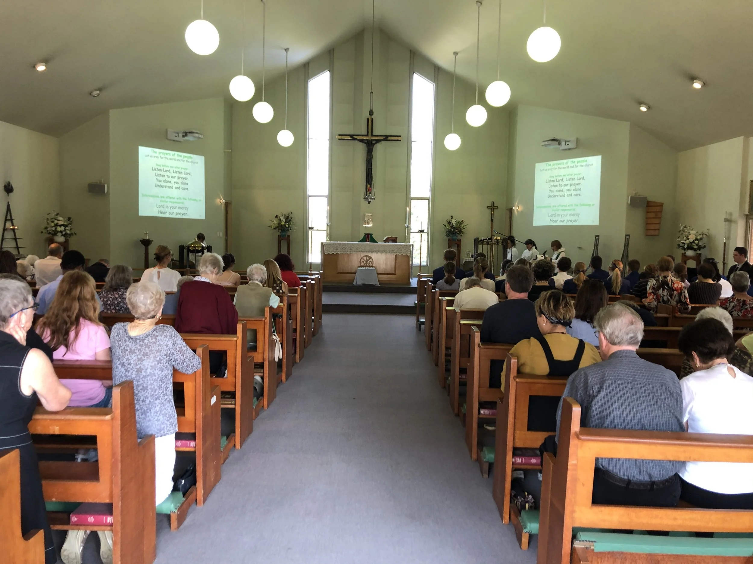 People seated in a church congregation during service, facing the altar with a large crucifix, stained glass windows, and two projection screens displaying song lyrics.