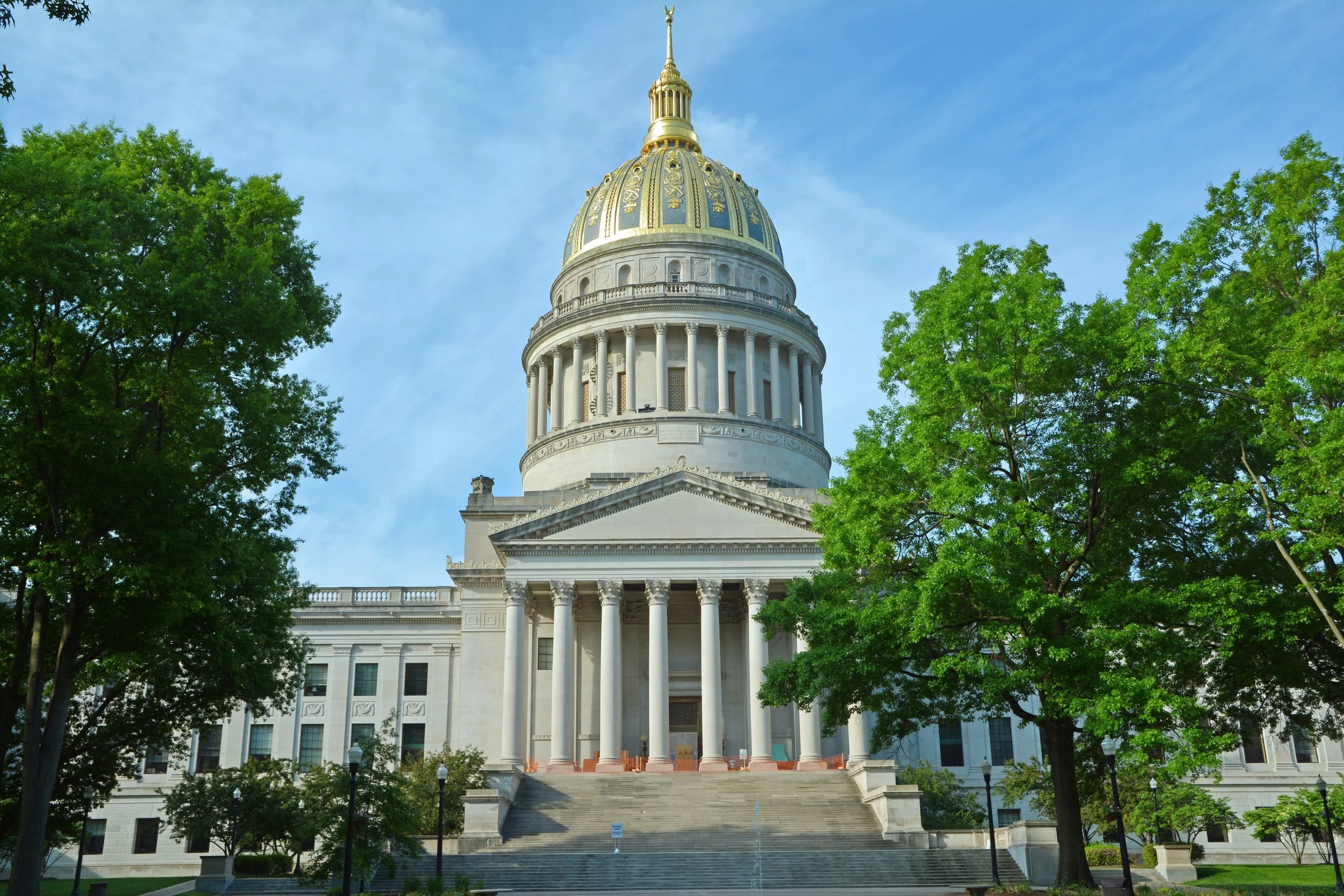 The image depicts the West Virginia State Capitol building, characterized by its neoclassical architecture with tall white columns and steps leading up to the entrance. 