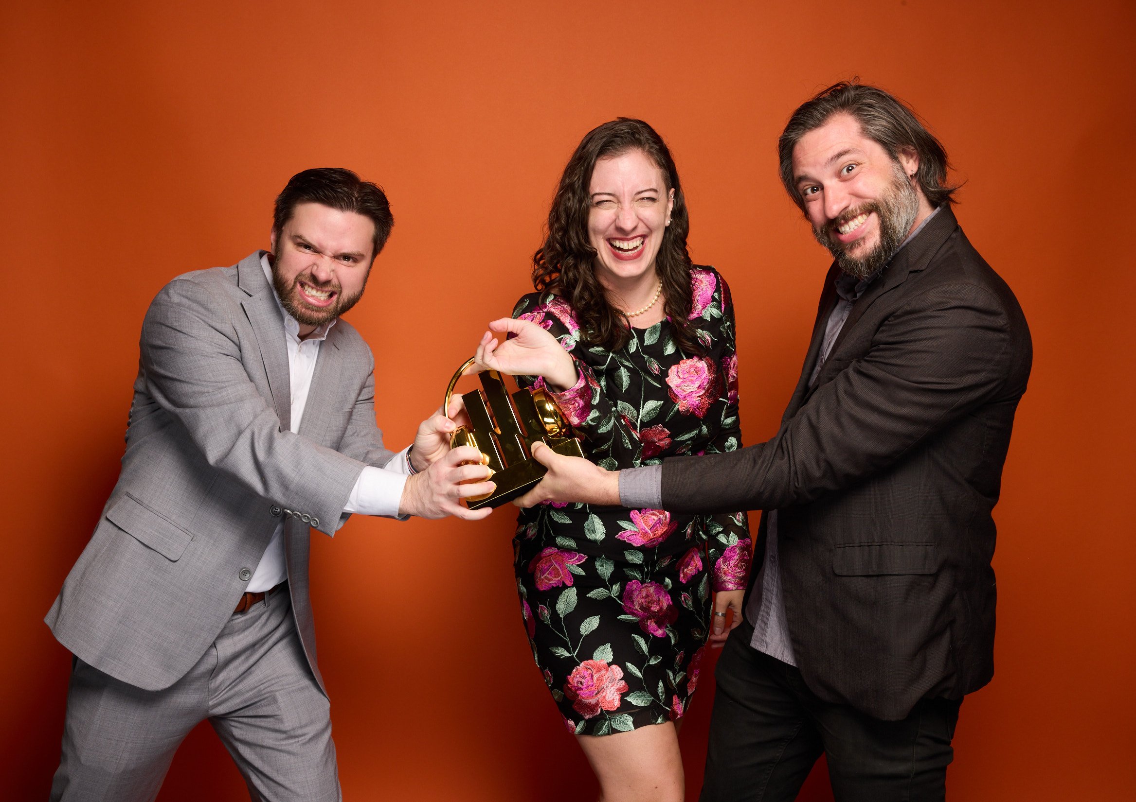 Three people, two men and one woman, smiling and posing against an orange background, holding a decorative trophy together.