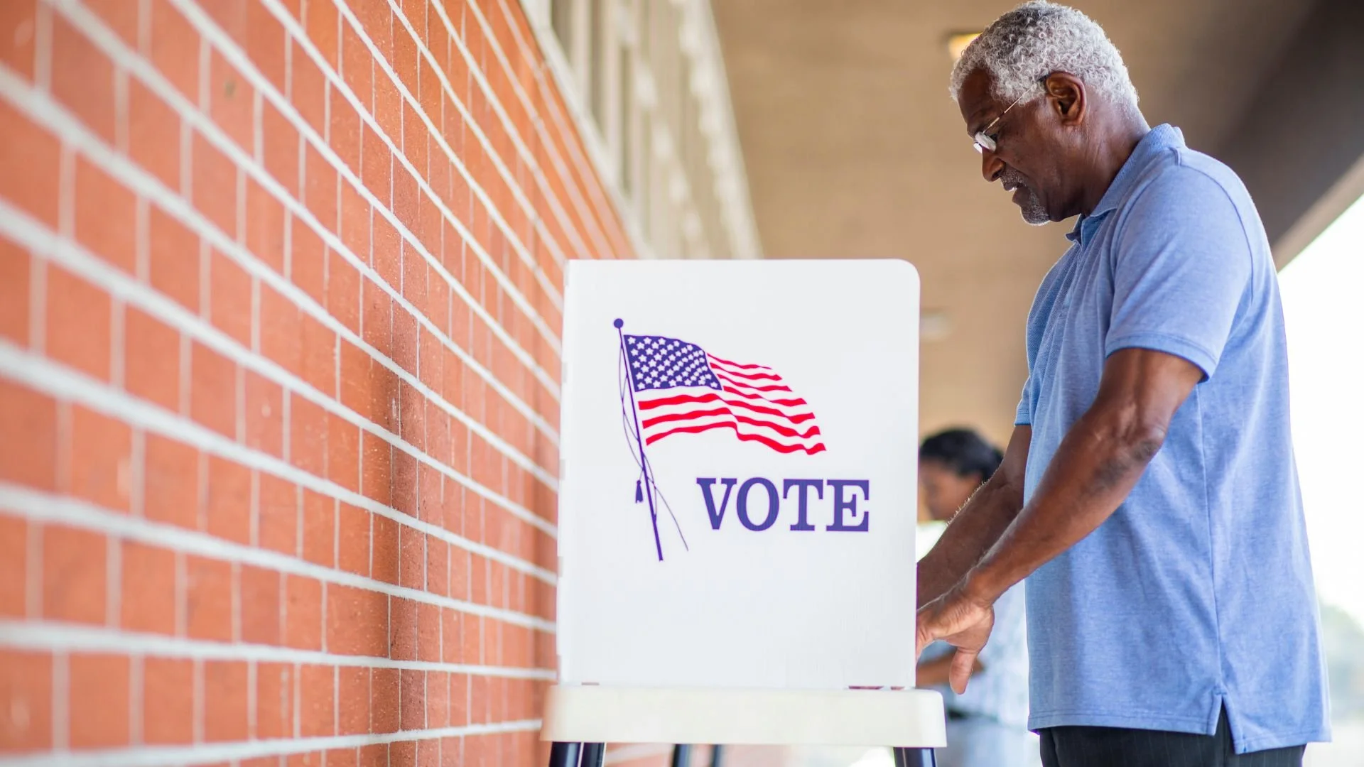 An older man voting at a polling station with a sign that features an American flag and the word 'VOTE' next to a red brick wall.