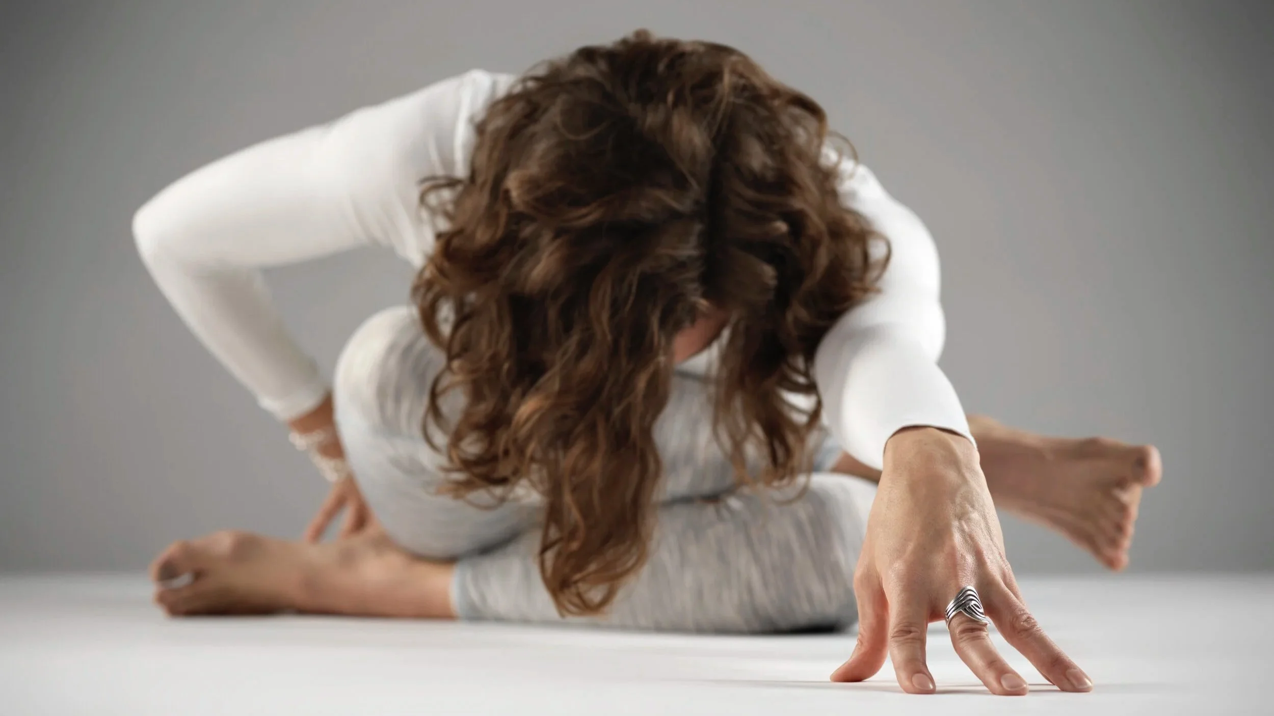 Woman practicing yoga on a gray background, sitting in a cross-legged position with her head bent forward, reaching out with her right hand, and wearing a white long-sleeve shirt and light-colored pants.