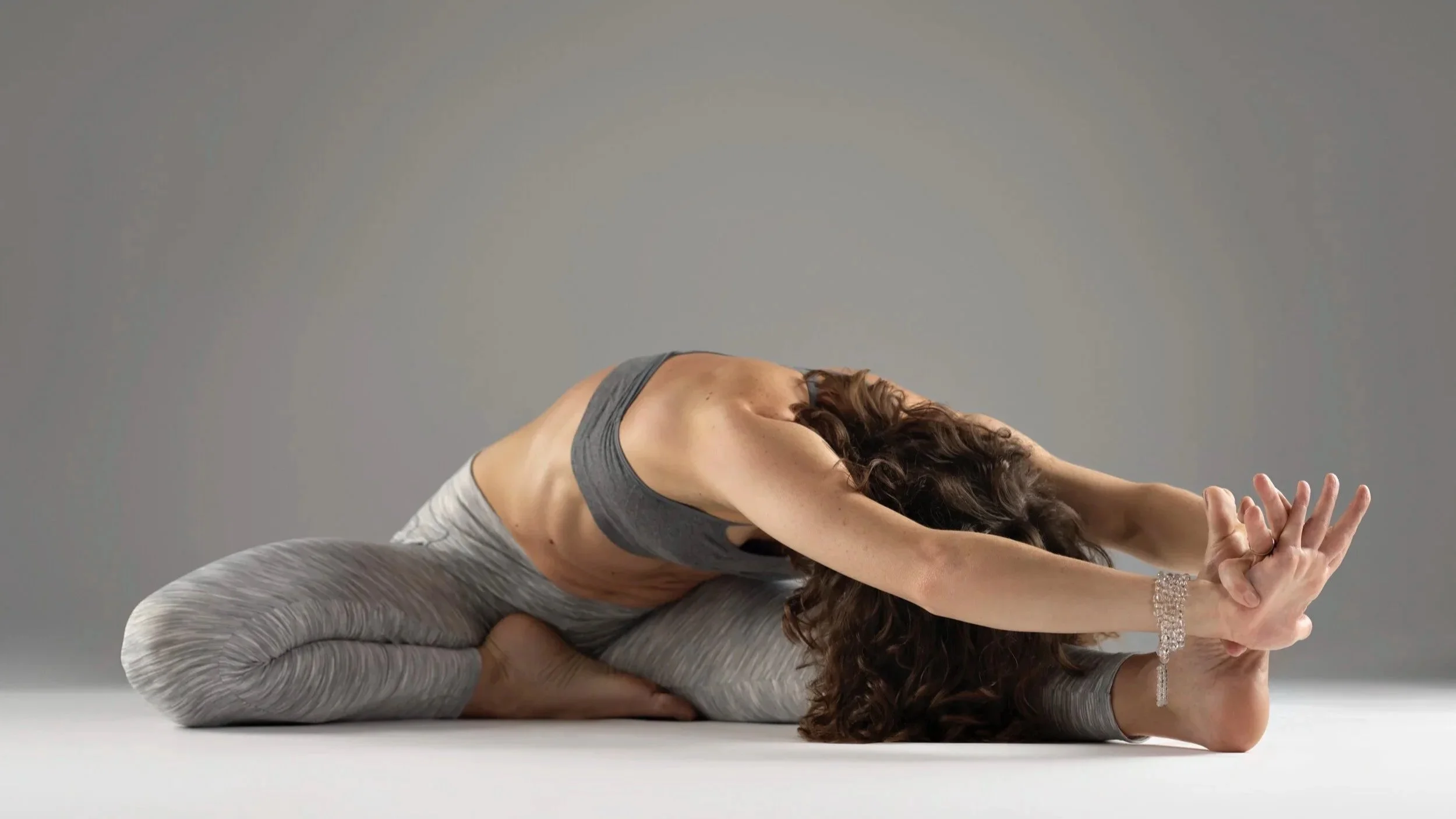 A woman practicing yoga on the floor, stretching her arms forward and holding her foot with her hands.