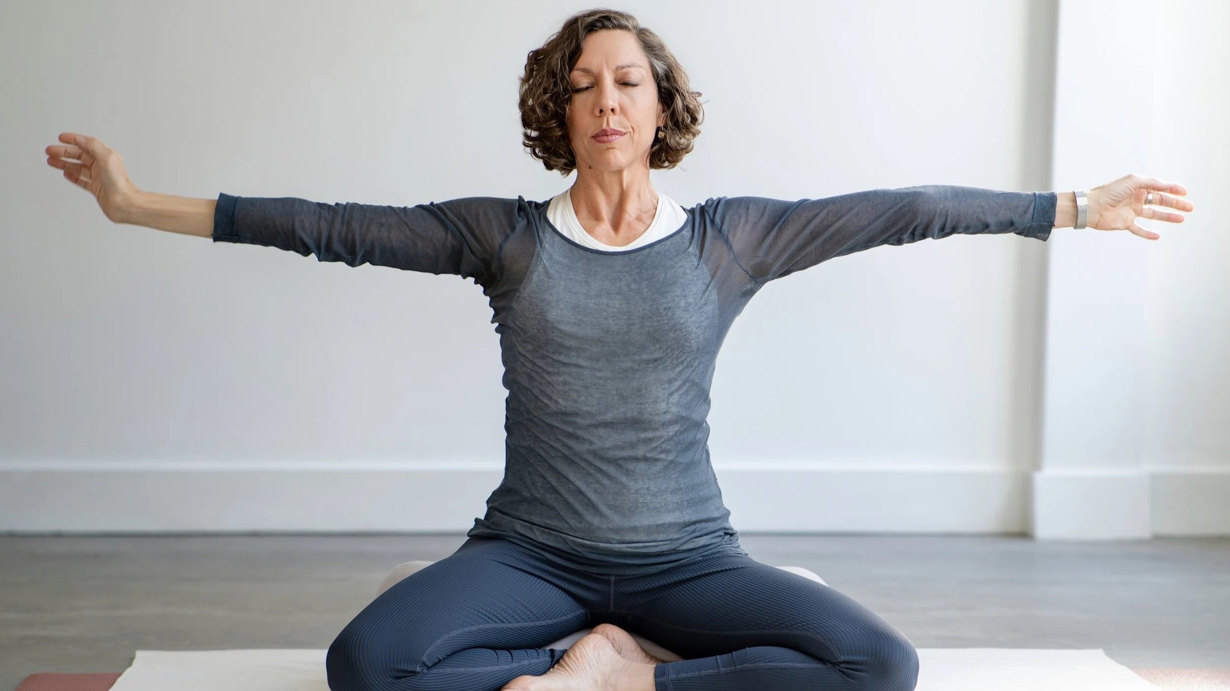 A woman practicing yoga indoors, sitting cross-legged on a mat with arms extended outward, eyes closed, and a peaceful expression.